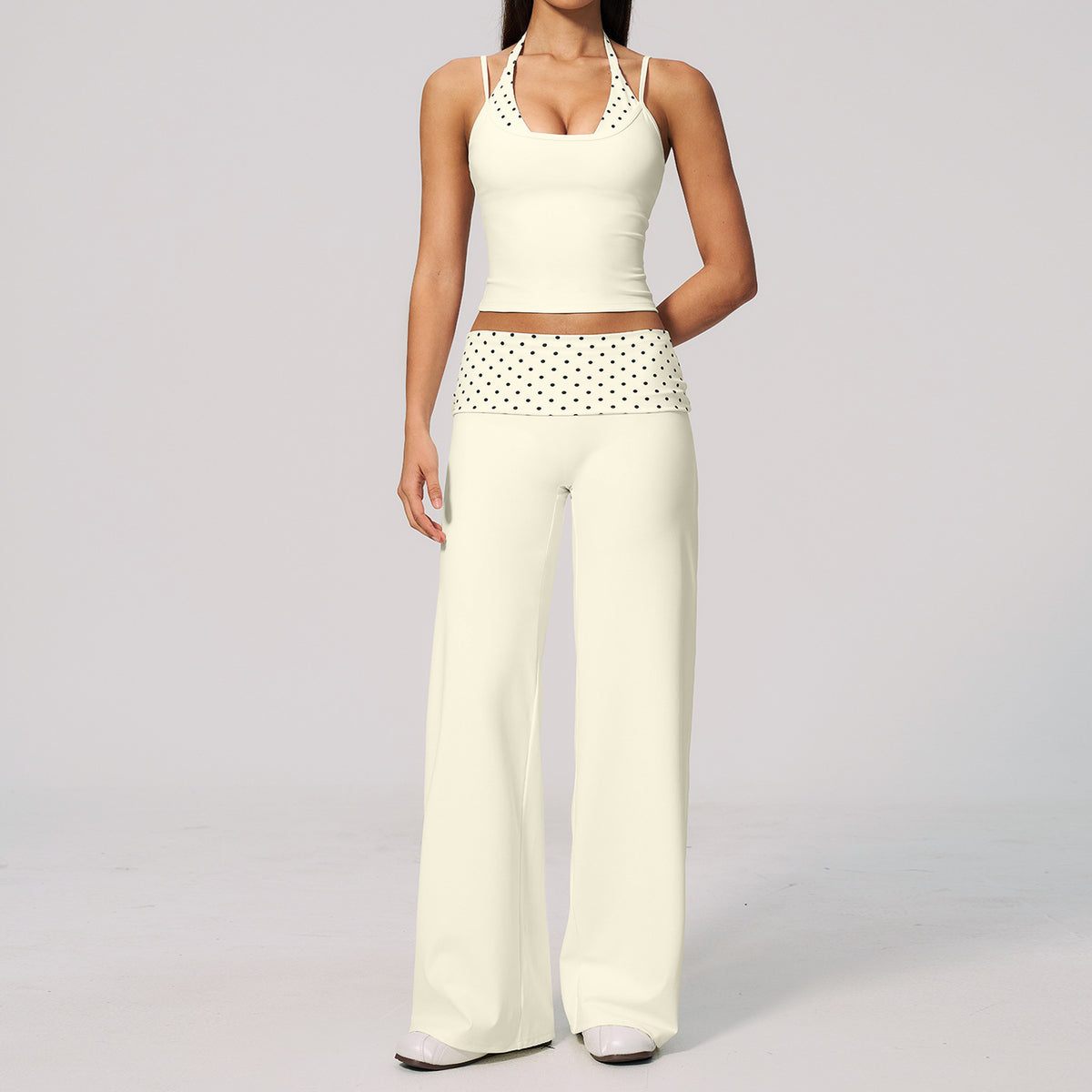 A model wearing a cream-colored two-piece lounge set consisting of a halter crop top with crochet detailing and matching wide-leg pants with a polka-dot waistband, set against a plain light gray background.